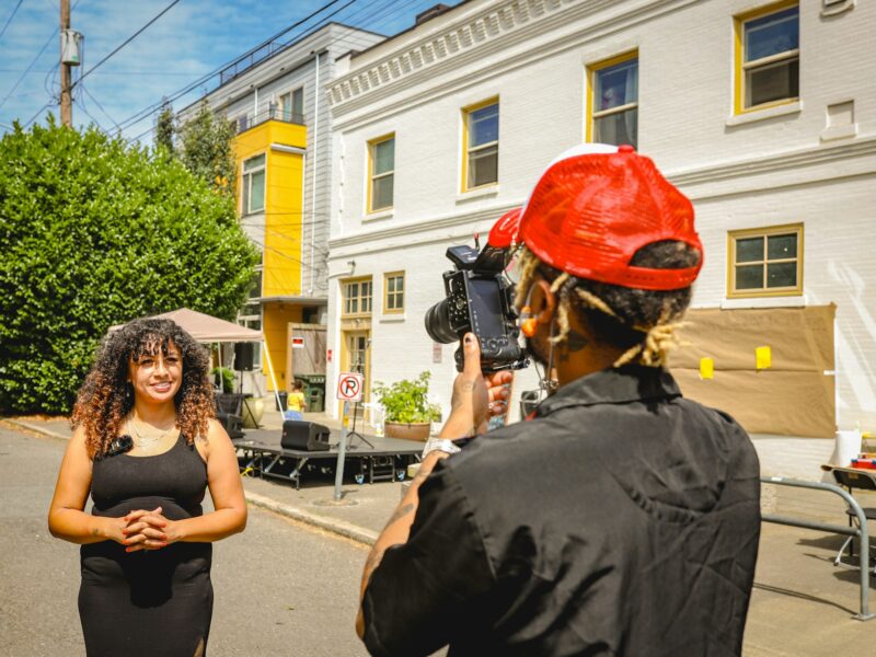 A camera operator filming a woman being interviewed on a neighborhood street
